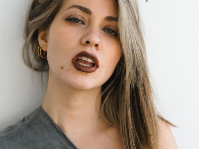 JuliannaXXX-hot has brown lipstick, slightly open mouth, small moles on her cheek, long blonde hair, and gold hoop earrings, wearing a grey shirt.