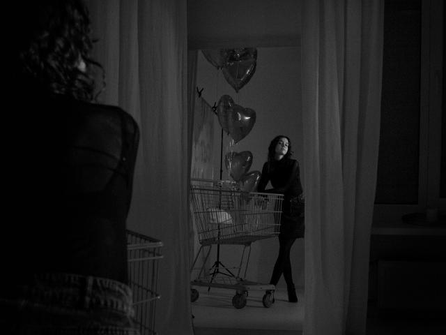 LettyCurly with curly hair, dark clothes, and black mask, standing in front of a shopping cart with heart balloons, framed by curtains, in a black-and-white shot.