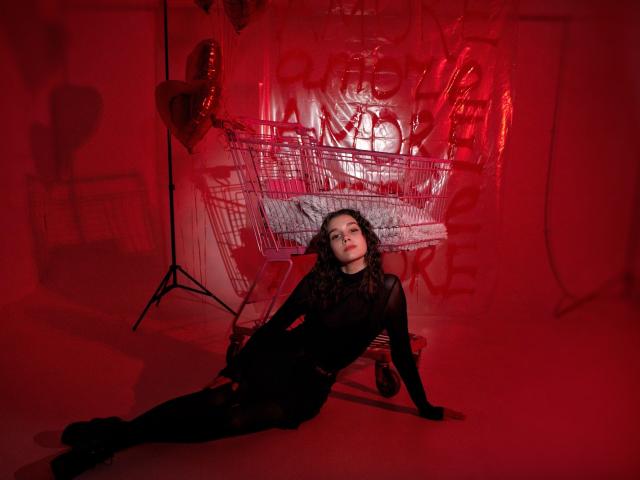 LettyCurly, with dark curly hair, wearing a black sheer dress, is seated on the floor, legs stretched out, in a red-lit room, with a heart balloon and shopping cart.
