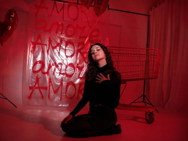 LettyCurly, with curly dark hair, kneels in red light wearing a black long-sleeve top, hand on chest, looking up, next to a wire shopping cart.