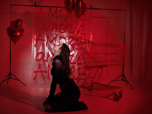 LettyCurly, kneeling in black lingerie, with curly hair, facing a red-lit shopping cart and heart balloons, in a red-washed room. Her ass is prominent, back to the camera.
