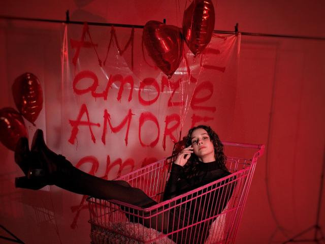 LettyCurly, avec ses cheveux noirs bouclés, vêtue de noir, est assise dans un chariot de supermarché rose, les jambes en l'air, portant des bottes noires, tenant des lunettes, des ballons rouges, éclairage rouge, séduisante, fond rouge foncé..