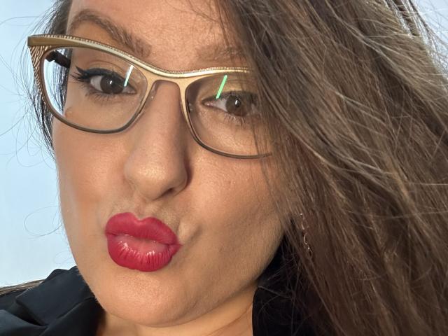 BadAngel with red lipstick, brown eyes, and glasses, pouting slightly. She has long brown hair and a black shirt. Her tits are not visible, just her face.