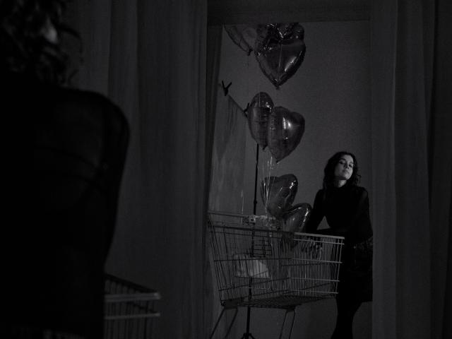 Black-and-white photo of LettyCurly with curly hair, standing near a metal shopping cart, holding shiny heart-shaped balloons, wearing a dark dress, looking up. Silhouette of another person.