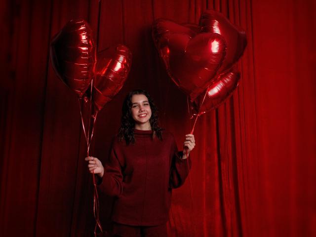 LettyCurly, avec ses cheveux noirs bouclés, portant un pull rouge, tenant des ballons en forme de cœur rouges brillants, souriante, debout devant un rideau rouge, ayant l'air mignonne et joyeuse..