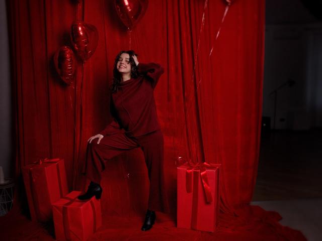 LettyCurly with curly brown hair, red outfit, and black shoes, smiling, leaning against red curtain, surrounded by red balloons and gift boxes. Red hearts on balloons.