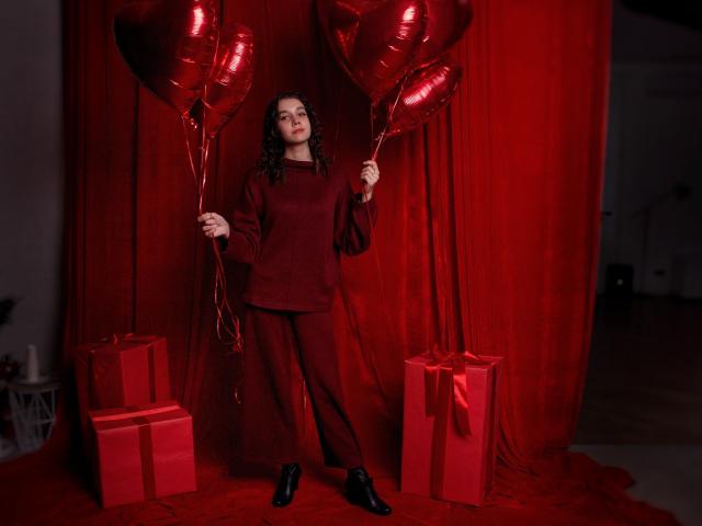 LettyCurly with curly brown hair, wearing a red dress and boots, holding shiny red heart balloons, standing in front of red gift boxes. Red curtain backdrop.