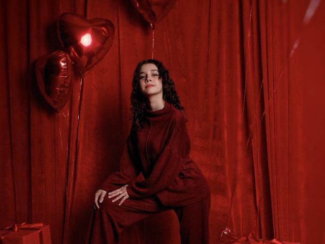 LettyCurly with curly dark hair, wearing a red sweater and pants, sitting against red curtains with heart balloons, looking up with a slight smile, hands on knee.