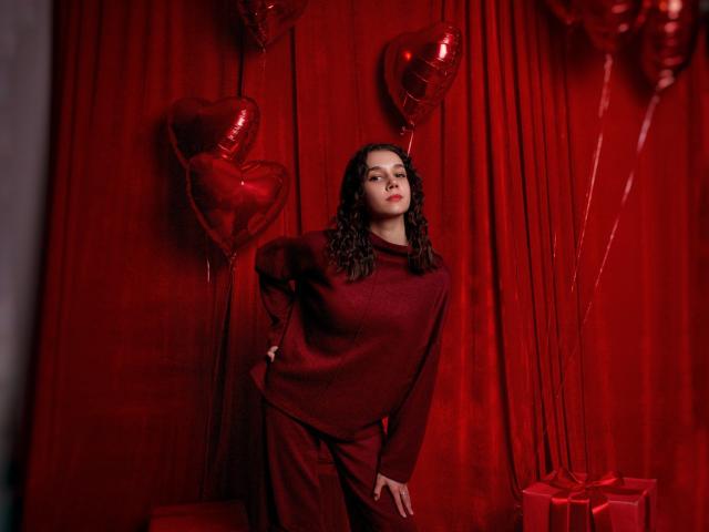LettyCurly with curly dark hair, wearing a red sweater and pants, stands against a red curtain with heart-shaped balloons, hand on hip, looking confident. Red gifts on floor.