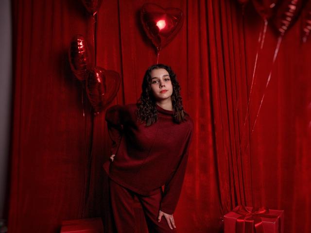 LettyCurly with curly brown hair, wearing a red sweater and pants, leaning against red curtains with heart balloons and gifts, looking at the camera with a slight smile.