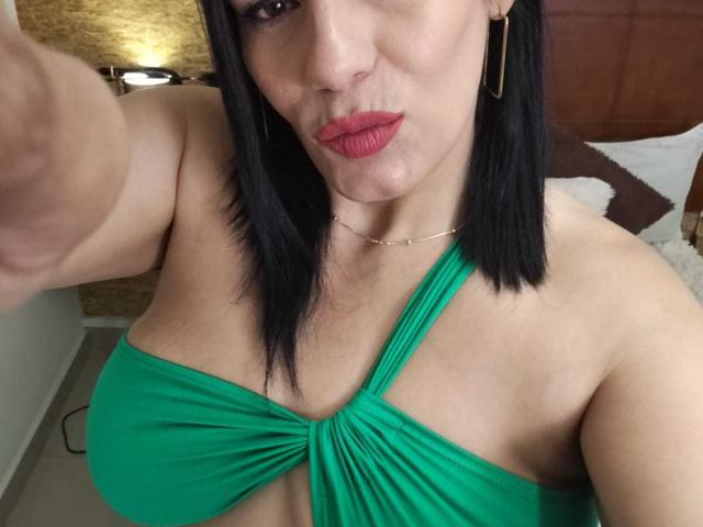 LizaRussell with black hair, green halter dress showing her large tits, red lipstick, gold earrings, and a necklace, taking a selfie, pouting her lips.