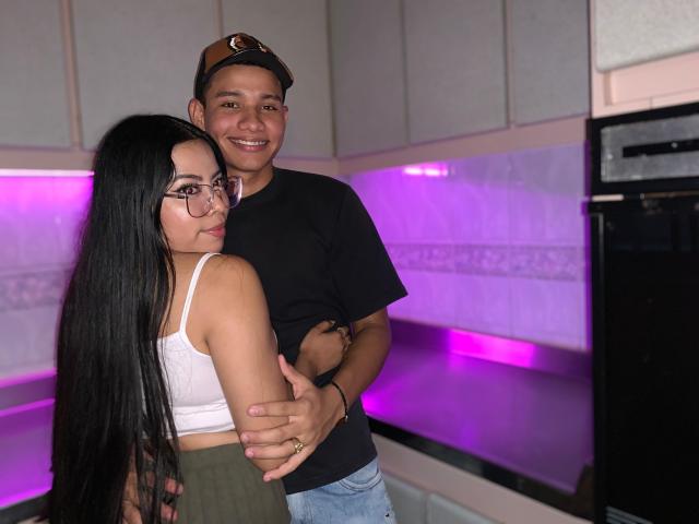SaraAndMoreno with long black hair, glasses, white tank top, green skirt, standing in front of SaraAndMoreno, smiling, black t-shirt, blue jeans, hand on her ass, purple lighting.