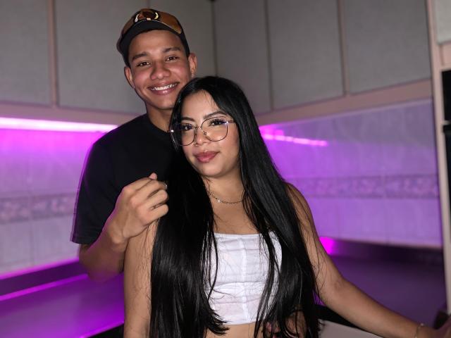 SaraAndMoreno with long black hair, glasses, white tube top, and small tits, standing in front of SaraAndMoreno with a black shirt, brown cap, and smiling.