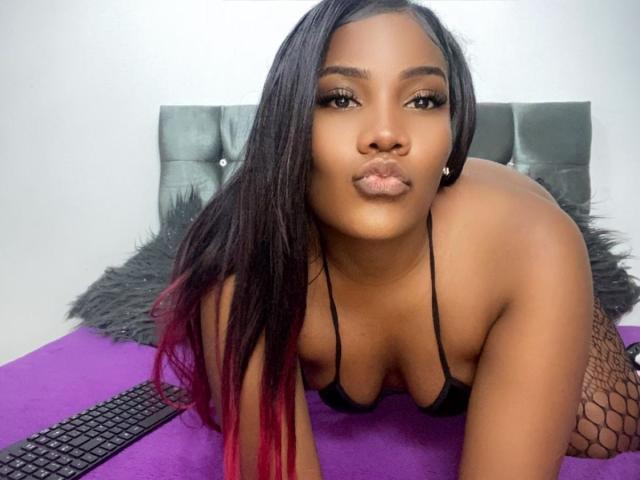 SamiiaSmith with dark skin, long black hair with red tips, pouty lips, black bikini top, fishnet legs, on purple bed, gray headboard, keyboard in foreground.