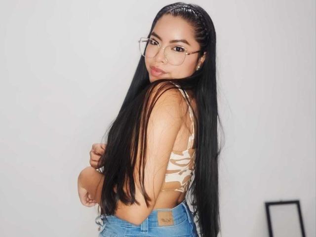 SaraMoreno with long black hair, glasses, tan camo crop top, and blue jeans, looking over shoulder, showing small ass and slim back, in a simple indoor setting.