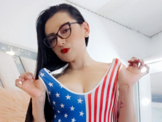 NataliaCamp with black glasses, red lipstick, and long black hair, wearing a sleeveless American flag dress, pulling up the top to show her tits.