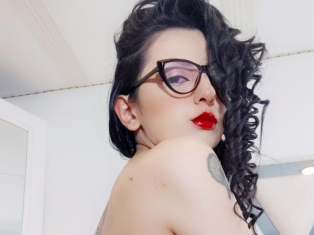 NataliaCamp with curly black hair, red lipstick, and glasses, showing her bare shoulder with a tattoo, looking over her shoulder, pale skin, and a sexy, confident expression.