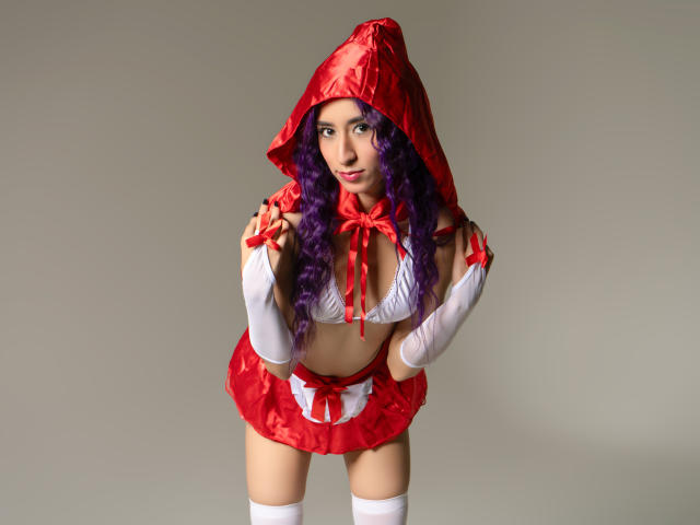 Julia, com cabelo cacheado roxo, vestindo um conjunto vermelho com capuz, um sutiã branco, uma saia vermelha, meias brancas até a coxa e uma fita vermelha, curvada para frente, mostrando os seios, em uma foto de estúdio..