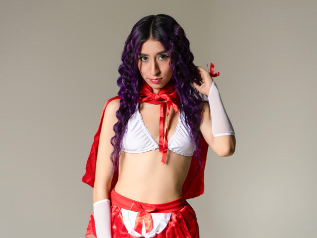 JuliaJobs with purple curly hair, wearing a red and white cosplay outfit, has her hand on her head, showing her small white bra and red ribbon. Boobs, tits, red skirt.