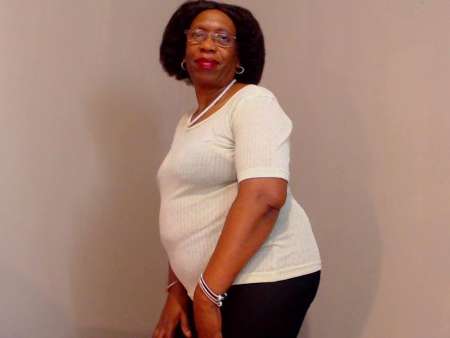 FreakyAssX, dark-skinned woman with short curly hair, glasses, white ribbed shirt, black pants, pink lipstick, standing, smiling, wearing white necklace, black bracelet.