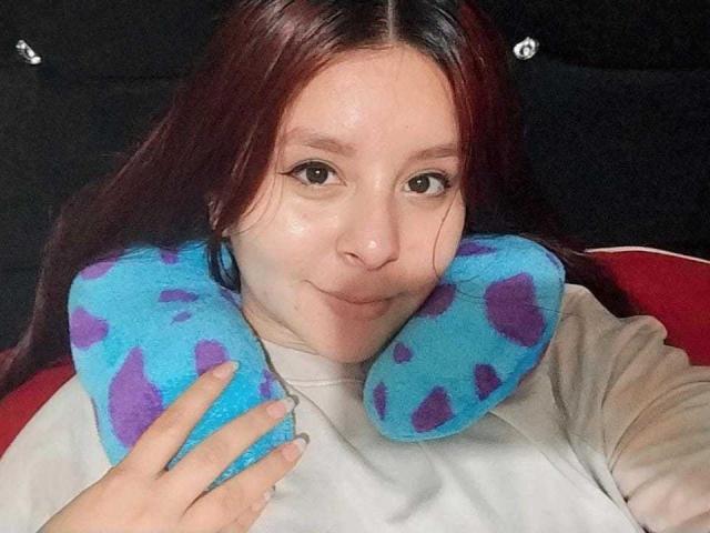 EmaJames with red hair, holding a blue and purple pillow against her shoulder, wearing a white shirt, smiling slightly. She's in a red chair.