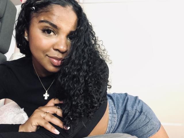 LisaMendez with curly black hair, black top, and blue shorts, sticking out tongue, wearing cross necklace, lying on bed, showing cute face and hand with rings.