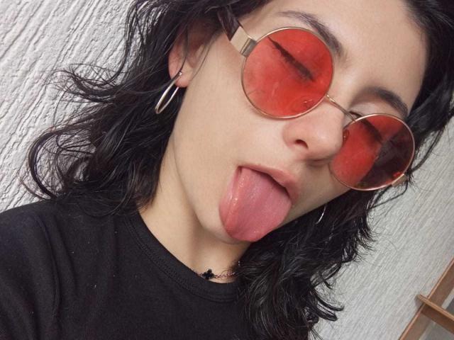 JeneKurz with black wavy hair, red-tinted round glasses, and tongue out, wearing a black shirt, hoop earrings, and a small black necklace.