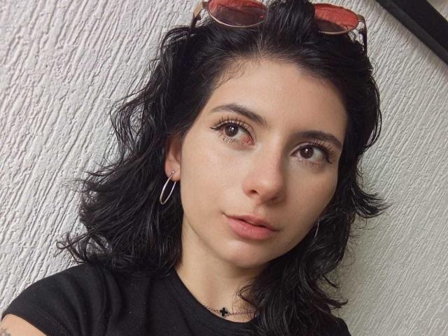 JeneKurz a les cheveux noirs ondulés, les yeux marrons, et porte une chemise noire avec des boucles d'oreilles cercles. Des lunettes de soleil reposent sur sa tête. Son visage est légèrement boudeur, avec une expression neutre..