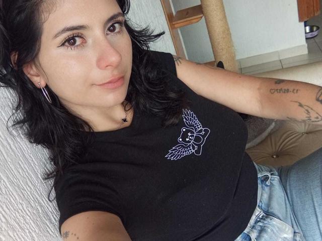 JeneKurz with black wavy hair, wearing a black tee showing a bird design, jeans, tattoos on arms, earrings, sitting, smiling, and showing small tits.