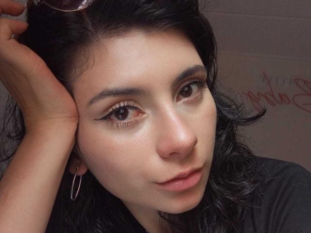 JeneKurz tem cabelos escuros e ondulados, olhos castanhos com cílios grossos, e usa brincos de argola. Sua mão está no cabelo, mostrando uma maquiagem discreta e uma expressão relaxada..