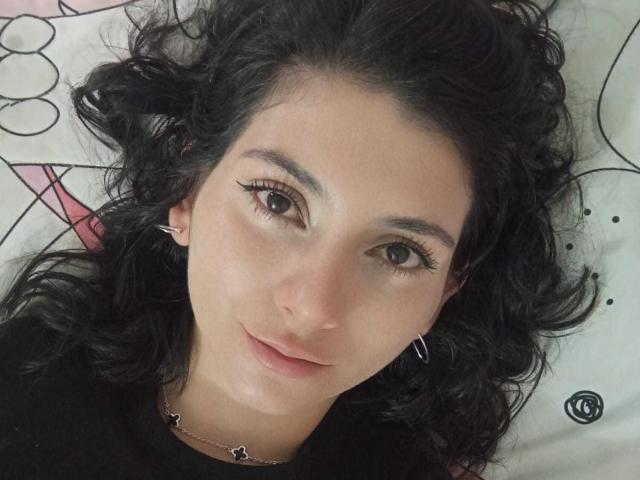 JeneKurz has curly black hair, brown eyes, and is wearing a black top with star earrings, lying on a patterned surface, showing a subtle smile.