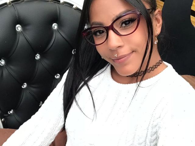 Amberv has black hair, red glasses, a nose piercing, white sweater, choker, and earrings. Her ass is not visible, just her face and upper body.