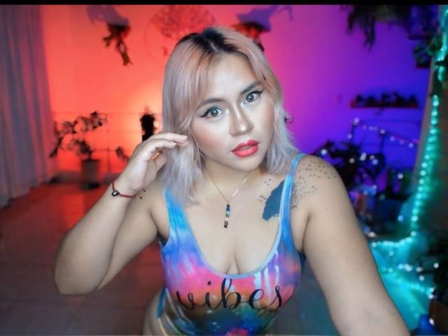 FairyNia with blonde hair, blue eyes, and red lipstick, wearing a tie-dye tank top, showing her tits, tattoos on her shoulder, and a necklace, in a colorful room.