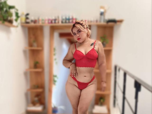 FairyNia vestindo um conjunto de lingerie vermelha de renda, com a mão na cintura, mostrando os seios e o bumbum, cabelo loiro, batom vermelho, tatuagem no ombro direito, fundo borrado..