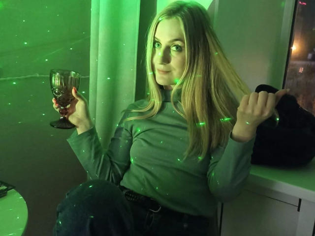 Kristinakokietka, con i capelli verdi e luminosi, tiene in mano un bicchiere, indossa una maglietta verde, jeans scuri e una borsa nera, ed è seduta vicino a una finestra. Particelle verdi luminose la circondano..