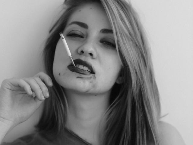 JuliannaXXX-hot with a cigarette in her mouth, dark lipstick, and a slightly droopy expression, holding her chin with her hand. Black and white, messy face.