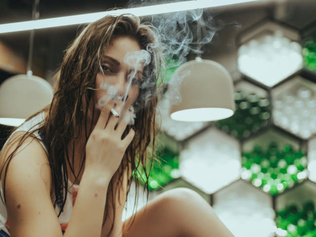 JuliannaXXX-hot with wet brown hair, smoking a cigarette, her hand near her mouth, with smoke curling around her face, wearing a white tank top, in a modern setting.