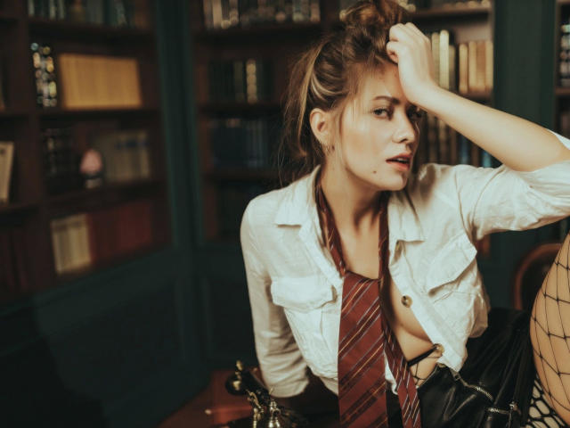JuliannaXXX-hot, with dirty blonde hair, sits in a library, wearing an open white shirt, red striped tie, and black fishnet stockings, hand on head, looking to the side.