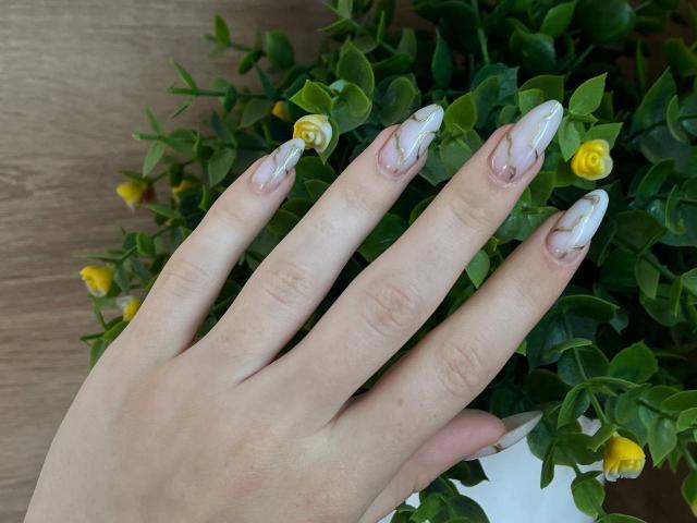 A mão de Valissia, com unhas longas e esmaltadas em um branco marmorizado, repousa delicadamente sobre uma planta verde com rosas amarelas. Suas unhas são elegantes, e sua pele é clara..