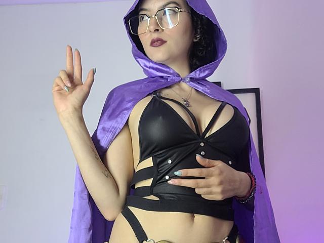 GabrielaMoore in a purple hood, black strappy bra, showing her tits, with a hand up and a finger gesture, wearing glasses and a ring.
