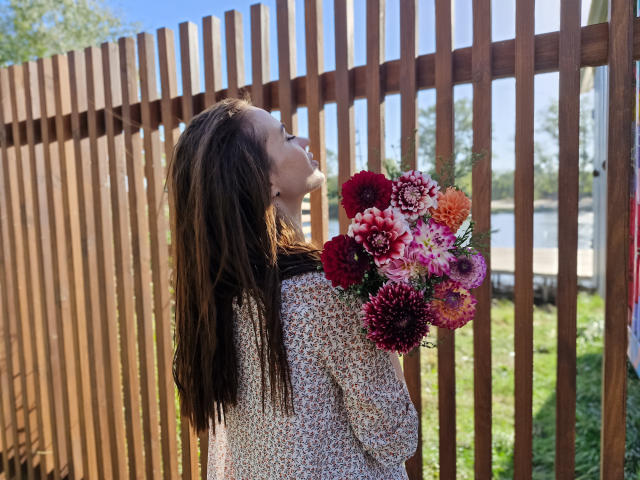 KaraCriegs with long brown hair, wearing a floral dress, holding a vibrant bouquet of red, pink, and orange flowers, standing in front of a wooden fence.