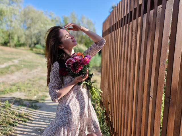 KaraCriegs steht an einem Holzzaun, die Augen geschlossen, und hält einen Strauß aus rosa und roten Blumen, sie trägt ein gemustertes Kleid, die Sonne scheint ihr ins Gesicht, und darunter befindet sich ein grasbewachsener Pfad..