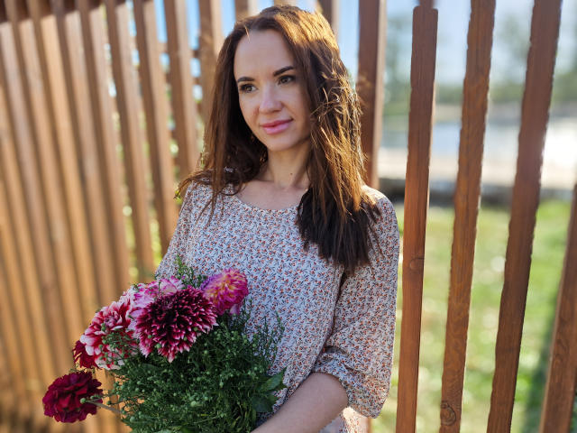 KaraCriegs with long brown hair, floral dress, holding pink and red chrysanthemums, standing by wooden fence, smiling, sunlight on her face, green foliage in background.