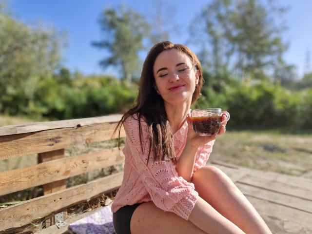 KaraCriegs met lang bruin haar, een roze gebreide top en zwarte shorts, zittend op een houten bankje, thee drinkend, met gesloten ogen, lachend, met zonlicht op haar gezicht, een glas vasthoudend in haar rechterhand..