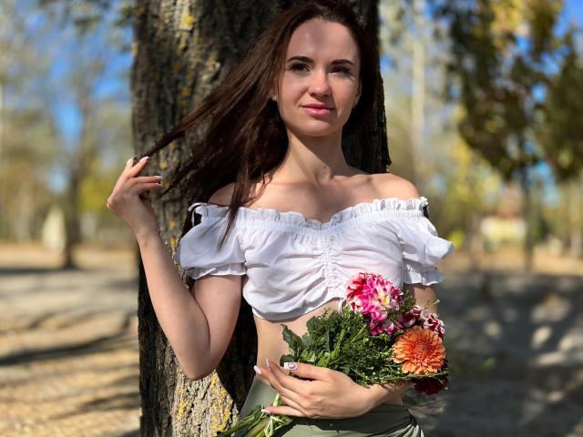 KaraCriegs with long brown hair, off-shoulder white top, holding colorful flowers, standing by a tree, sunlight on her face, slightly smiling, holding hair with right hand.