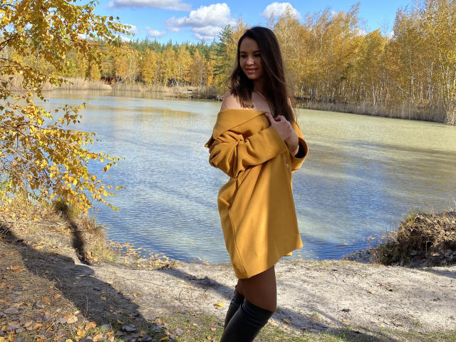 KaraCriegs with long brown hair, off-shoulder yellow sweater, black knee-high boots, standing by a clear lake, autumn trees, and blue sky in the background, looking at the camera.