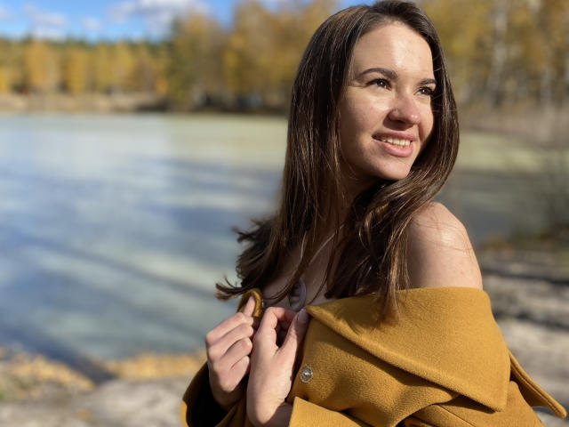 KaraCriegs mit braunen Haaren, lächelnd, hält einen senfgelben Mantel von ihren Schultern, wobei ihr entblößtes Oberteil sichtbar ist, steht an einem klaren, bläulich schimmernden See, im Hintergrund Herbstbäume..