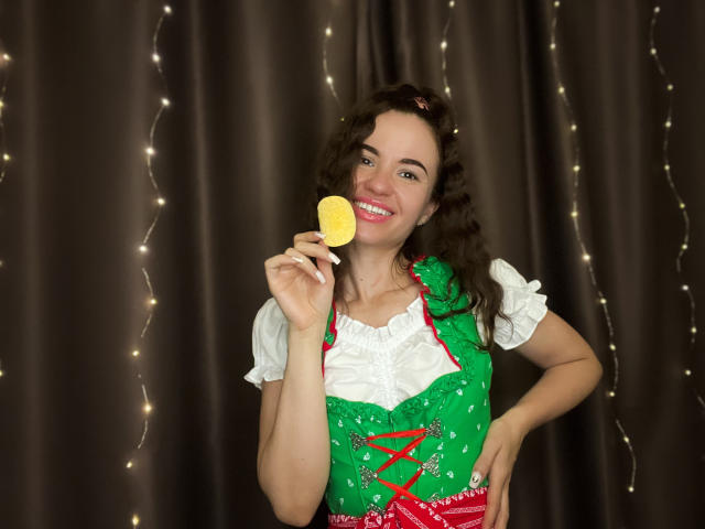 KaraCriegs with curly brown hair, smiling, holding a yellow lollipop, wearing a green and white Bavarian-style dress, standing, hands on hips, fairy lights in background.