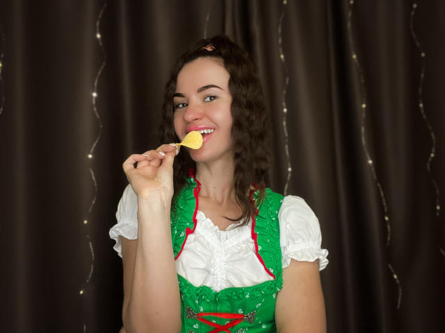 KaraCriegs with curly brown hair, wearing a green and white dress, smiling and sucking on a yellow lollipop, holding it with her right hand.