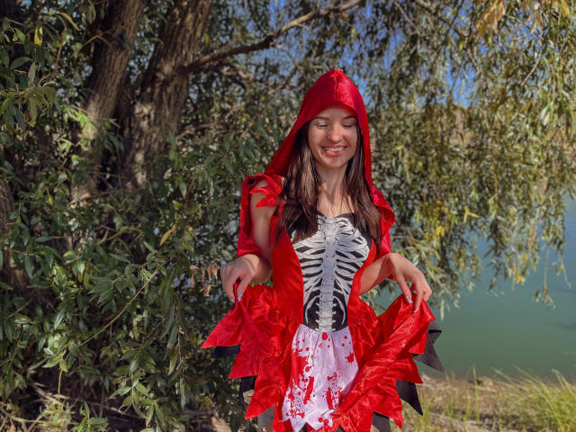 KaraCriegs, gekleidet in ein rotes Vampir-Kostüm mit Kapuze, einem schwarz-weiß gestreiften Korsett und einem roten Rüschenrock, lächelt, hält ihren Rock fest und zeigt ihre Brüste..